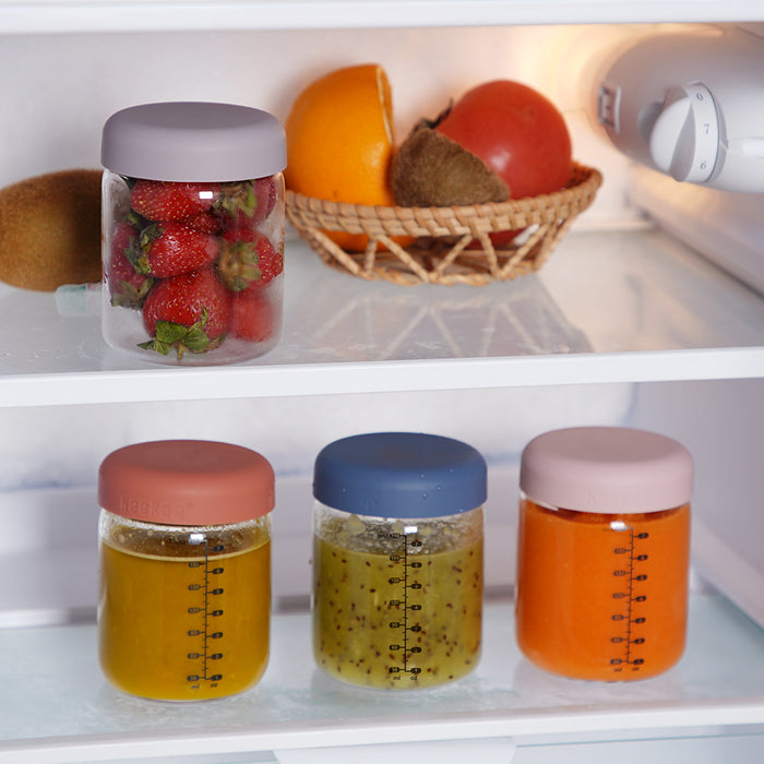The Haakaa Glass Baby Food Storage Jar Set neatly organizes a jar of strawberries, a basket with oranges and kiwi, and three vibrant jars of homemade purees on the bottom fridge shelf.