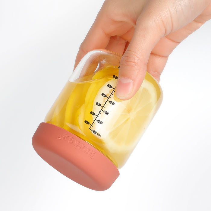 A hand holds a Haakaa Glass Baby Food Storage Jar with a red silicone lid, filled with lemon slices. The jar has measurement markings, ideal for homemade meals or baby food storage. The background is white.