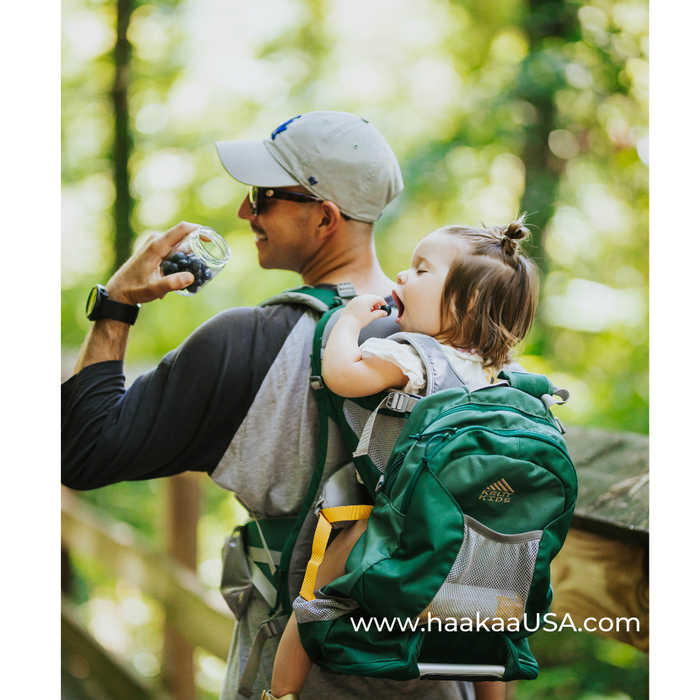 A man wearing a cap and sunglasses carries a toddler in a baby backpack carrier while holding a jar. The child, with a bow in her hair, is biting on the backpack's strap. They are outdoors with blurred trees in the background.