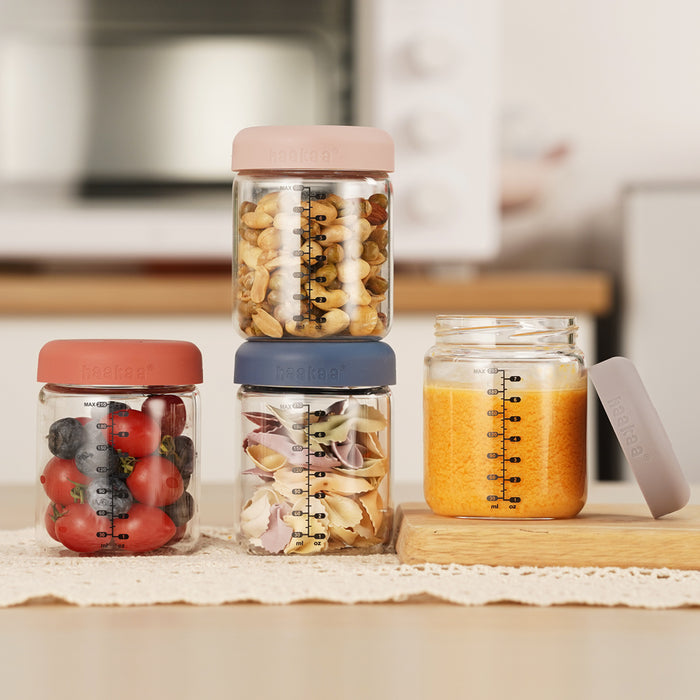The Haakaa Glass Baby Food Storage Jar Set includes four jars with colorful lids. Two are stacked with nuts, berries, and cherry tomatoes; a third holds garlic cloves, and the fourth has orange juice. Complete with measurement markings, one jar features its lid removed.