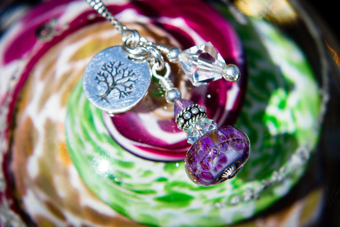 Close-up of a cremains infused glass bead necklace showing a purple bead, crystal accent and a tree engraved silver charm.