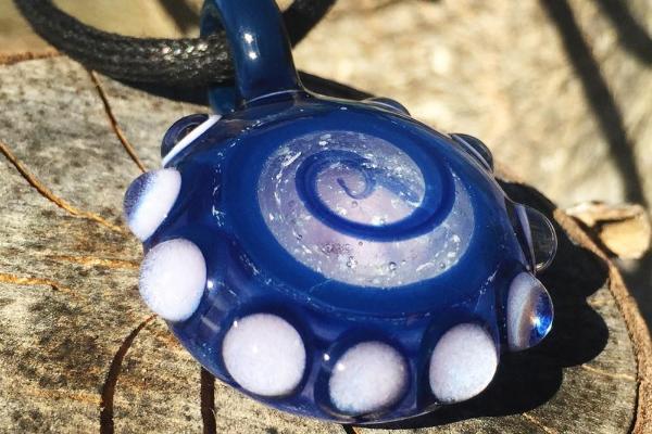 Blue glass bubble keepsake pendant with white edge dots and a swirl of cremation ash on the front, hanging from a cord.