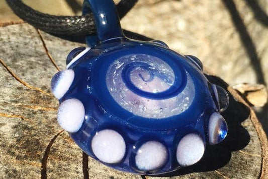 Blue glass bubble keepsake pendant with white edge dots and a swirl of cremation ash on the front, hanging from a cord.
