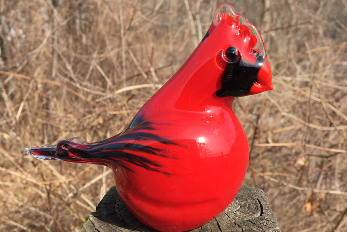 Glossy red glass cardinal cremation keepsake perched on a weathered stump outdoors.