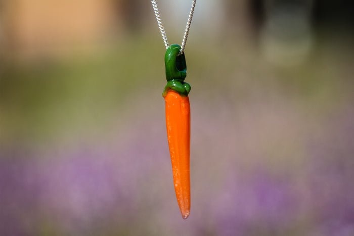 Orange glass carrot pendant with cremation ash and green top on a silver chain against a purple blurred background