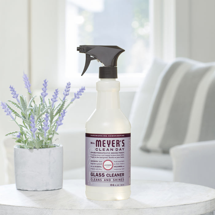 A Mrs. Meyers Glass Cleaner, free of ammonia, sits on a table beside a lavender potted plant. In the background, blurred white couch cushions complete a serene setting, adding charm and clarity to reflective surfaces.