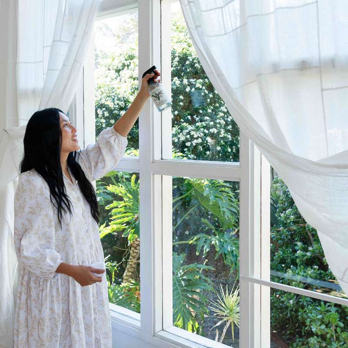 A woman in a light floral dress sprays Koala Ecos Natural Glass Cleaner on a large window in a bright room. She smiles amid sheer white curtains, with the fresh scent of Australian essential oils filling the air, while lush green garden plants are visible outside.