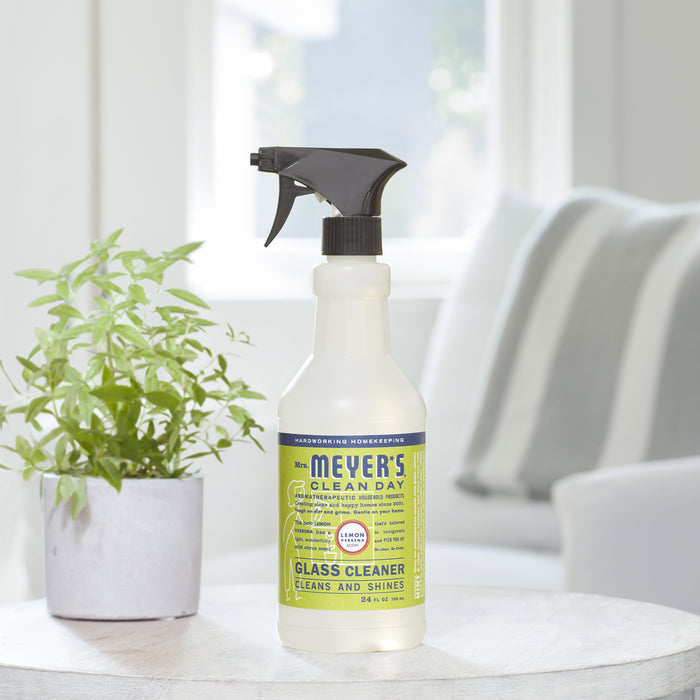 A spray bottle of Mrs. Meyers Glass Cleaner, ideal for reflective surfaces and ammonia-free, sits on a white table indoors. Beside it is a potted green plant, with a softly lit white couch and bright window in the background.