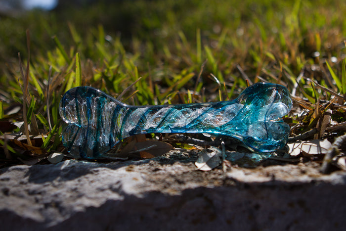 Teal and turquoise glass dog bone sculpture resting on grass.