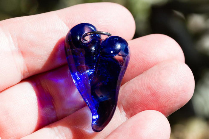 Cobalt blue glass heart cremation pendant with a metal loop held between fingers.