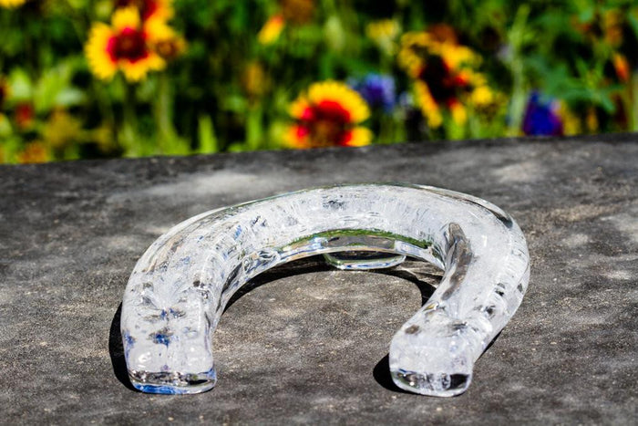 Clear glass horseshoe cremation sculpture on a dark stone surface with blurred flowers in the background.