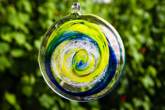 Two tone glass suncatcher infused with cremains featuring bright yellow and blue swirls hanging outdoors