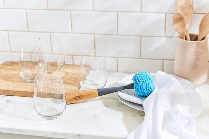 A kitchen scene exhibits four clear glasses, gleaming from Grove Co.s Glassware Cleaner Replacement Head, on a wooden board. A blue dish brush lies on a white towel next to wooden utensils in a holder, all beautifully arranged against a pristine white marble countertop and tiled backsplash.