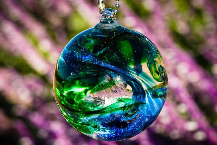 Glass cremation orb with green and blue swirls hanging against a blurred purple flower background