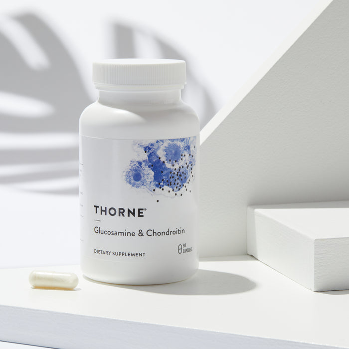 A white bottle of Thornes Glucosamine & Chondroitin dietary supplement, perfect for joint support, sits on a light surface next to a capsule. It features a blue abstract design and soft plant shadow in the background.