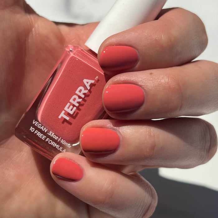 A hand holds a coral-colored Terra Beauty Bars bottle from the Good Vibes Kit, showcasing salon-quality shine. Nails painted in this vibrant vegan hue reflect light beautifully, with a white cap completing the look.