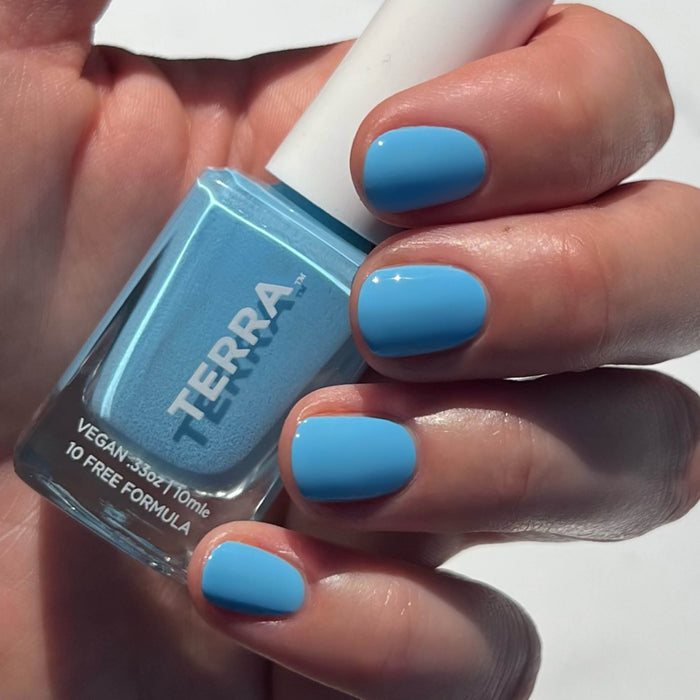 A hand with neatly manicured bright blue nails holds a bottle labeled Good Vibes Kit from Terra Beauty Bars. This salon-quality polish is vegan and 10-free, shining as a testament to superior nail care in the softly blurred background.