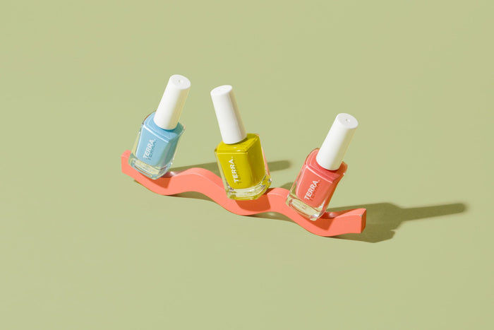 Three Terra Beauty Bars Good Vibes Kit nail polishes, featuring white caps and colors in pastel blue, yellow, and coral, sit on an orange wavy stand against a light green backdrop. This vegan polish promises salon quality, with shadows adding depth to the scene.