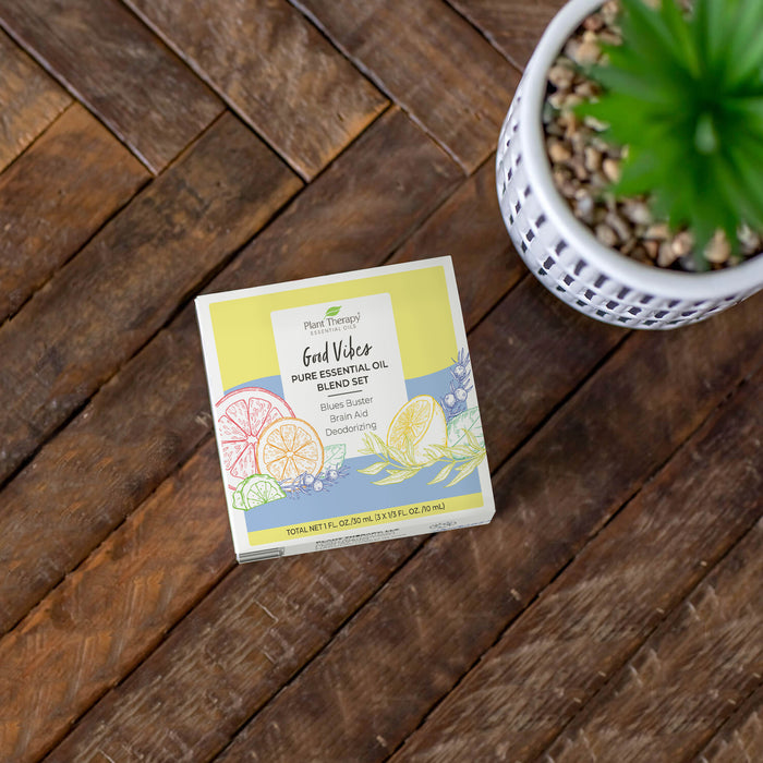 A box labeled Good Vibes Pure Essential Oil Blend Set by Plant Therapy is on a wooden surface, ideal for aromatherapy. It features colorful citrus slice illustrations, viewed from above with part of a white pot and green plant for an invigorating, natural deodorizing effect.