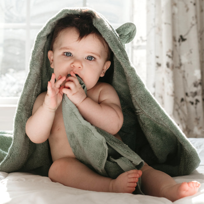 A baby with blue eyes and short dark hair sits on a white bed near a bright window, wrapped in a Natemia GOTS Certified, 100% Organic Cotton Hooded Baby Towel, featuring bear ears. The ultra-soft, absorbent green towel is held close to its mouth with both hands.