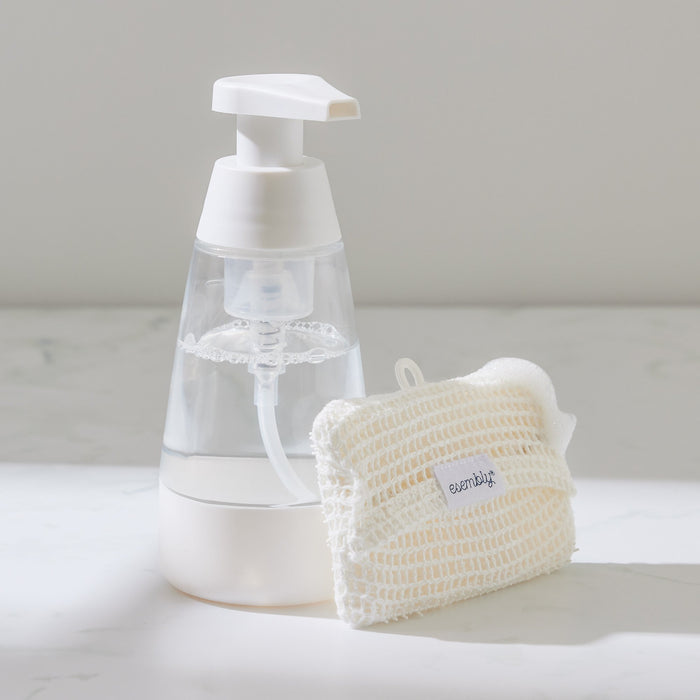 A clear foaming soap dispenser with a white pump is beside a woven beige pouch on a white surface, labeled everfit. The scene includes Esembly Babys GoTwo™ Kitchen Sponge Set, featuring a bacterial-resistant sponge, softly lit for a clean and minimalist look.