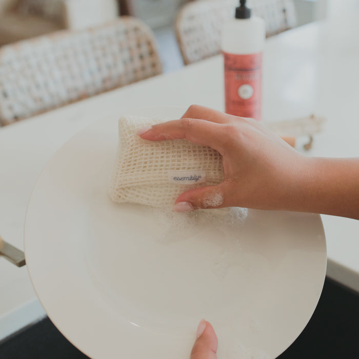 A person uses an Esembly Baby GoTwo™ Kitchen Sponge Set to wash a white plate, creating suds. A bottle of pink dish soap sits in the background on the countertop, and a wicker chair is partially visible.