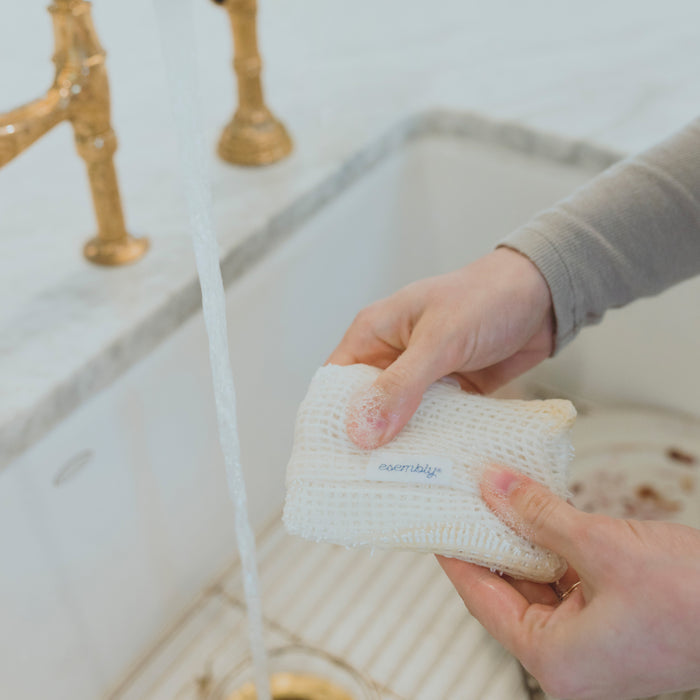 Hands using an Esembly Babys GoTwo™ Kitchen Sponge under running water from a gold faucet, with a marble sink and a few food scraps in the background.