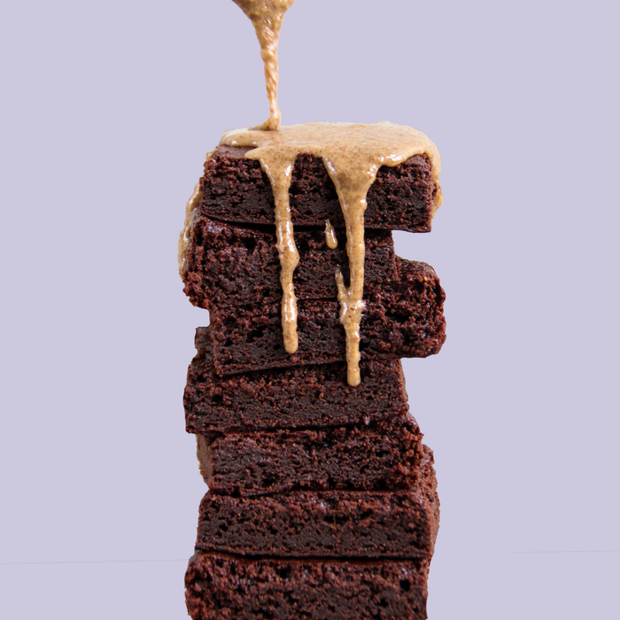 A stack of five decadent Stellar Eats Grain-Free Fudge Brownies, made with almond flour, sits against a light purple backdrop. A rich caramel sauce elegantly drizzles over the top and down the sides to create an irresistible picture of sweetness.