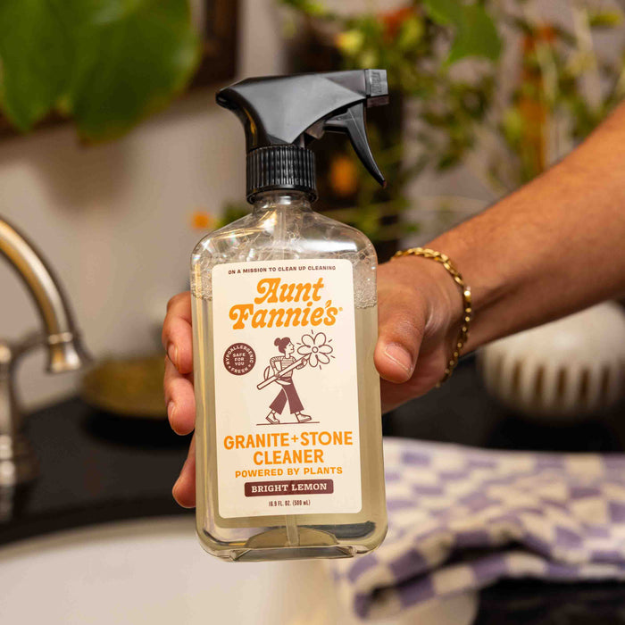 A hand holds Aunt Fannies Granite + Stone Cleaner, a natural cleaner labeled Bright Lemon, in a kitchen near a sink and a purple-checkered towel.