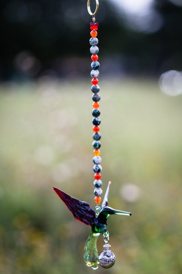 Vertical strand of orange and gray marbled beads with a green hummingbird charm and a silver keepsake urn hanging below