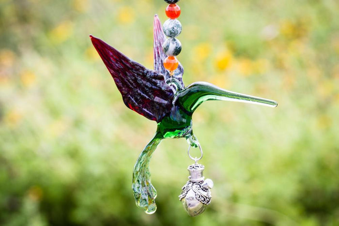 Green glass hummingbird with red wing outdoors connected to a beaded necklace and a decorative silver keepsake urn