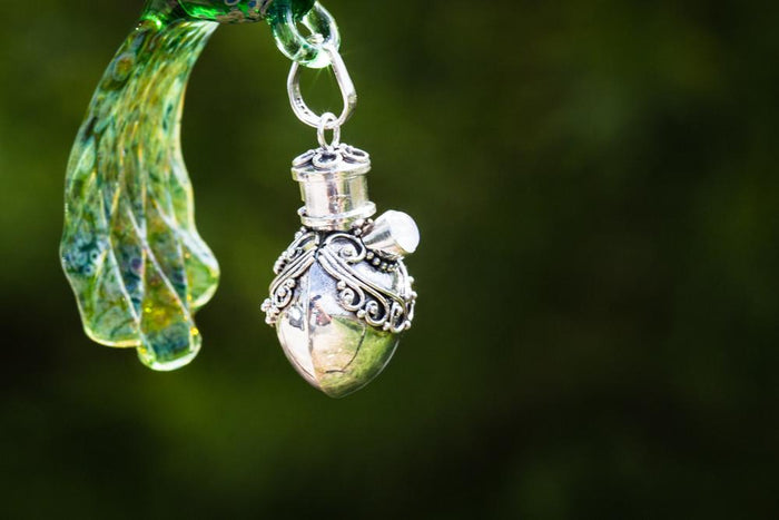 Close-up of ornate silver keepsake urn pendant with scrollwork and a green glass leaf accent