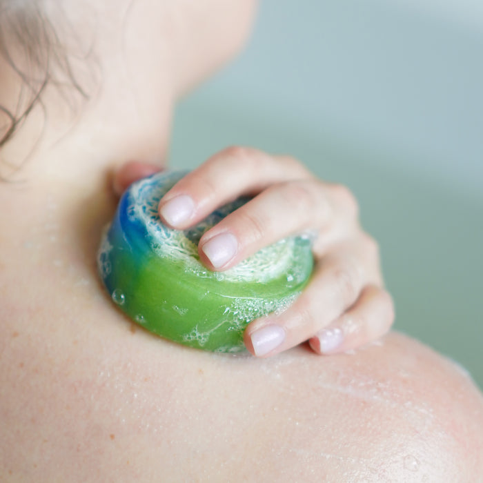 A person enjoys a soothing bath with Soapymania’s Magical Island Loofah, a vegan soap bar in blue and green. The suds glide over wet skin, creating an eco-friendly cleansing ritual that exudes cleanliness and pure tranquility.