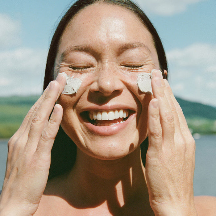A smiling person with closed eyes applies Ursa Major’s Green Slate Mineral Polish to their cheeks outside, with a blurred natural backdrop of sky and greenery.
