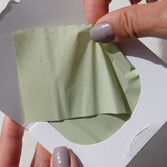 Close-up of hands peeling a green, speckled Green Tea Oil Control Paper infused with anti-inflammatory green tea extract from a white Ere Perez packaging envelope. The soft-textured paper is partially unfolded, casting gentle shadows in the light.
