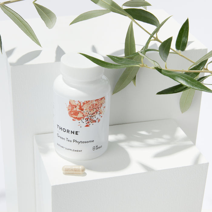 A white Green Tea Phytosome bottle from Thorne rests on a geometric white pedestal with two beige capsules nearby, suggesting antioxidant benefits. Olive branches arch above, enhancing the natural appeal of this fat-burning scene.