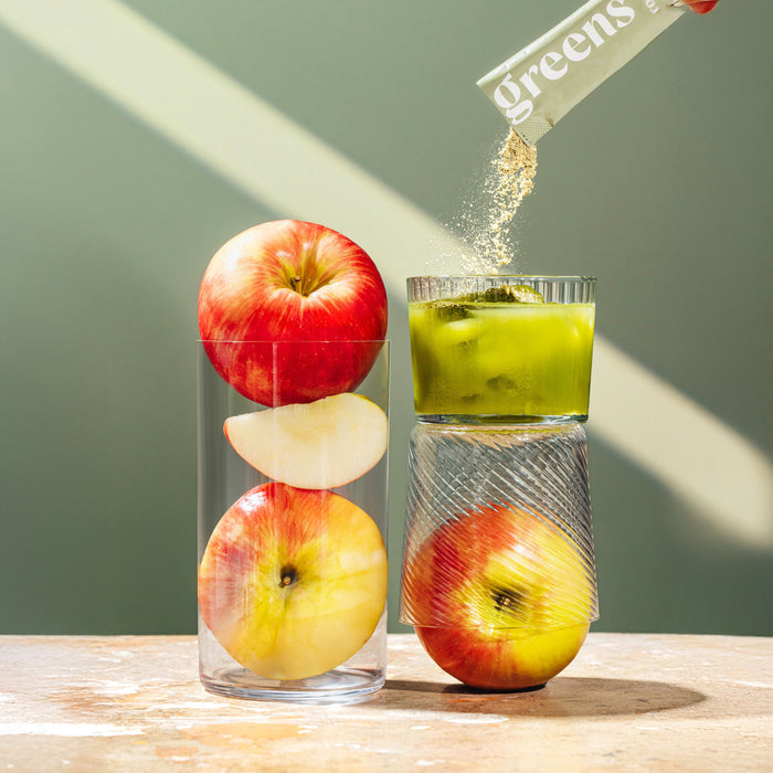 Two clear glasses are stacked; the left has whole and sliced apples, while the right contains green liquid and an apple as Rookie Wellness Greens + Superfood powder is poured in from a sachet.