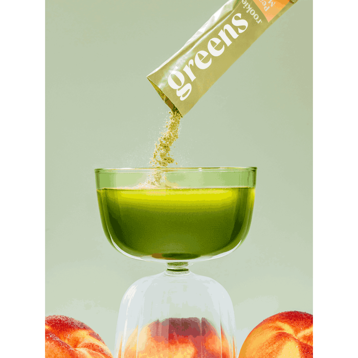 A glass of green liquid is shown as Rookie Wellness Greens + Superfood powder is poured in, promoting gut health. Two peaches are partly visible at the bottom on a light green background.