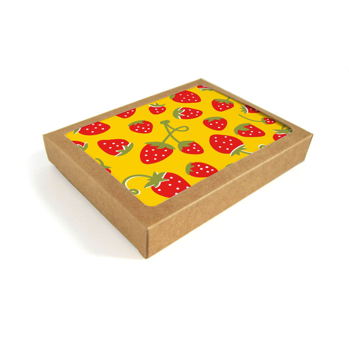 A brown recyclable cardboard box with a lid featuring original artwork of vibrant red strawberries on a bright yellow background, ideal for storing The Neighborgoods Greeting Cards, is placed diagonally against a pristine white backdrop.