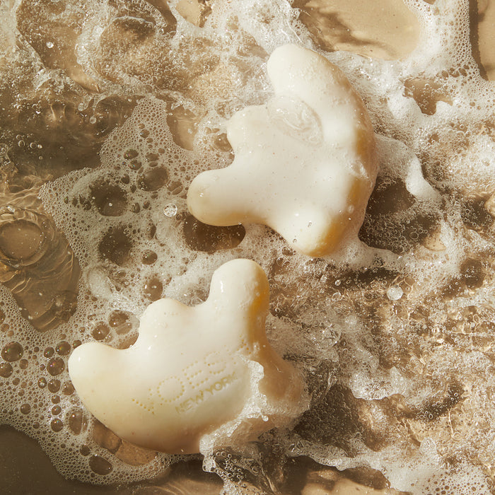 Two white, crescent-shaped Gua Sha Shampoo Bars by VOESH New York, featuring subtle brand engraving and finger indentations, rest on a beige sandy surface surrounded by foamy bubbles for a fresh and clean impression.