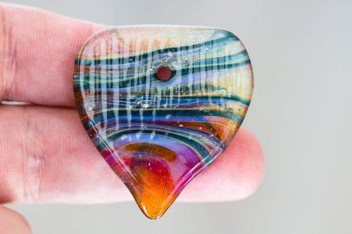 Rainbow swirled glass guitar pick infused with cremation ashes held between fingers