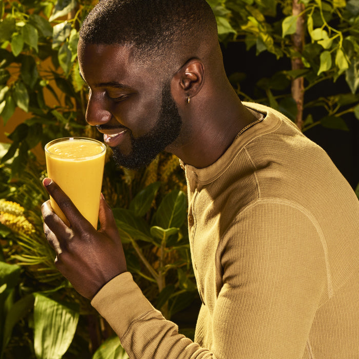 A man smiles while holding a glass of yellow Gut Bliss Organic Superfood Blend by Kuli Kuli Foods near his face, surrounded by lush green plants.