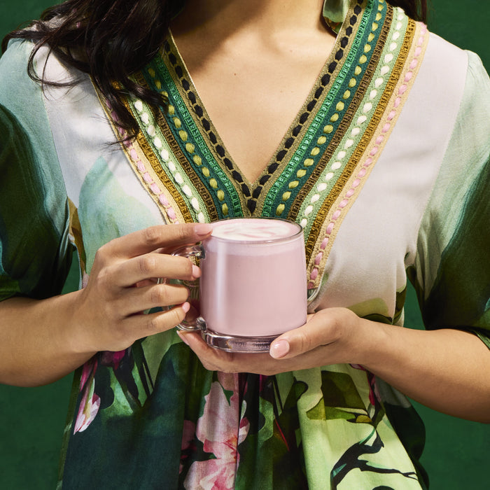 A woman in a colorful dress holds a clear glass mug of Kuli Kuli Foods Gut Health Organic Superfood Latte, filled with a pink hibiscus drink, standing against a green background. Only her torso and hands are visible.