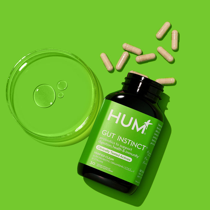 A black bottle of HUM Nutritions Gut Instinct Probiotics - For Digestive Health rests on a green backdrop, promoting digestion. Nearby capsules spill out, while to the left, a transparent container with droplets suggests nourishing your gut microbiome.