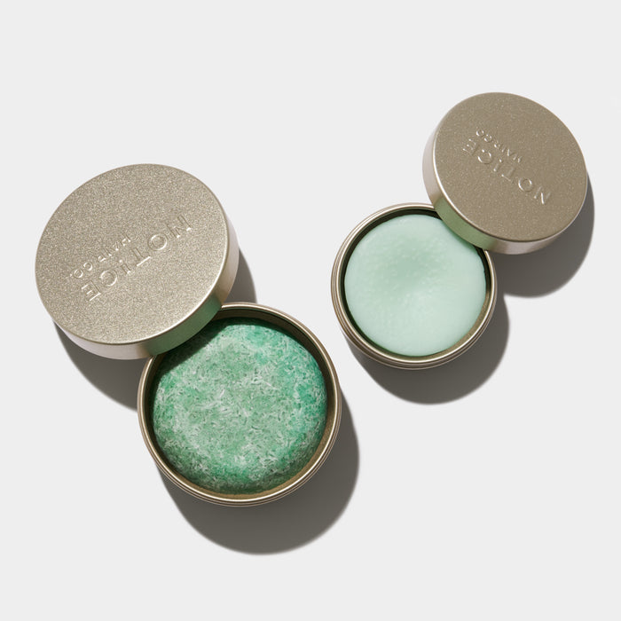 Two recyclable Hair Bar Travel Tins by NOT!CE Hair Co sit on a light background, lids ajar to reveal solid products: the left tin is textured and plastic-free with a green disc, and the right has a smooth, pale green disc. Both embody zero-waste elegance.