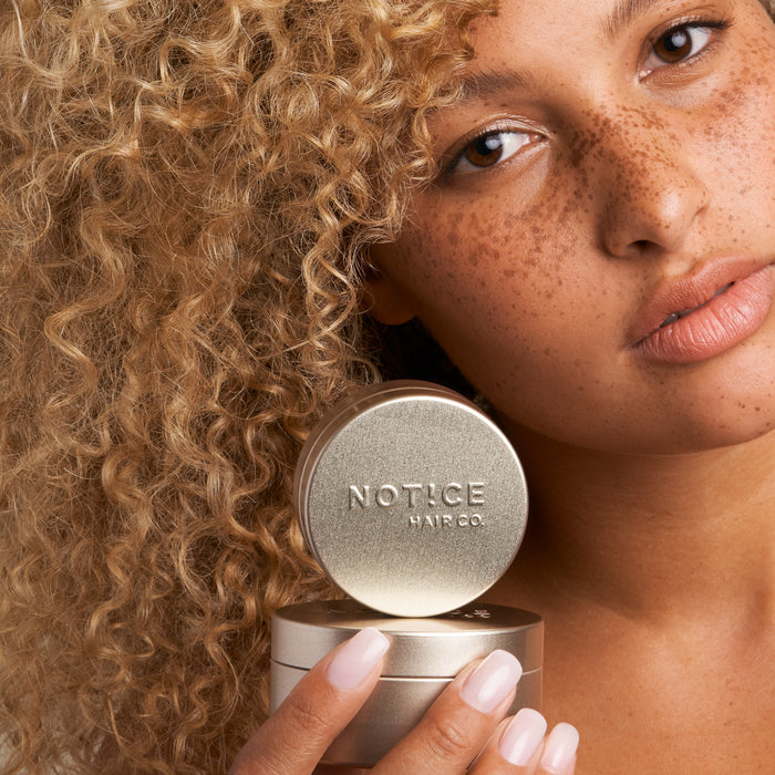 A freckled, curly-haired individual with nude lipstick holds a round metallic container of Hair Bar Travel Tins by NOT!CE Hair Co close to their face, highlighting its plastic-free design against a plain white background.