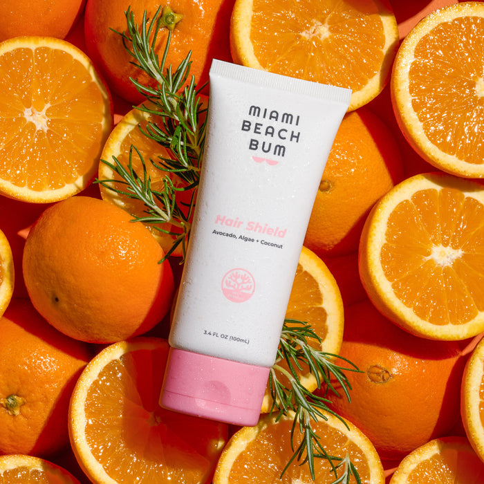 A tube of Hair Shield by Miami Beach Bum, a reef-friendly leave-in conditioner, lies among oranges and greenery. Its white body and pink cap pop against a bright backdrop, offering sun protection thats safe for color-treated hair.
