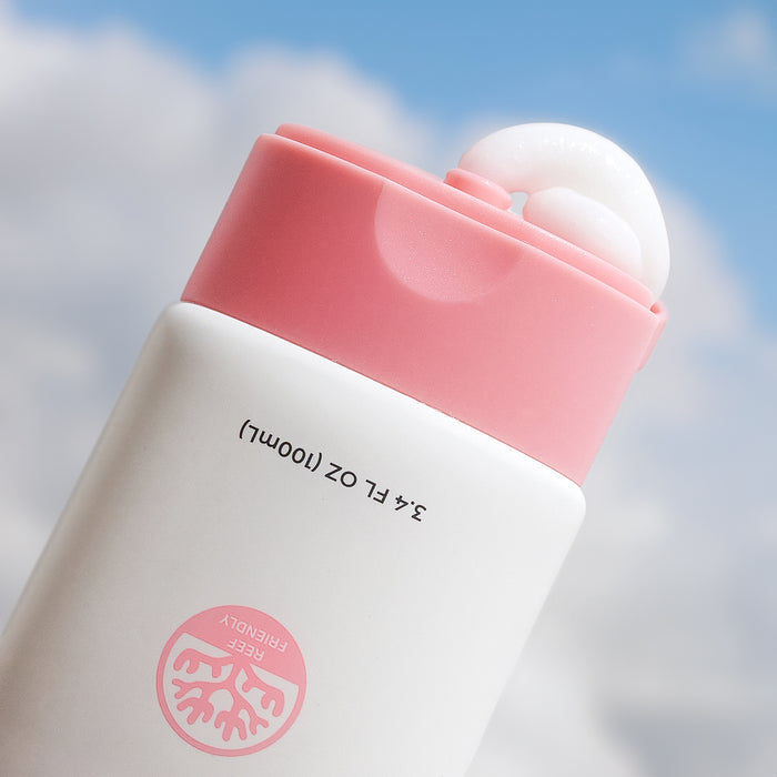 A white bottle of Hair Shield by Miami Beach Bum, capped in pink, leans against a cloudy sky. The open cap reveals some reef-friendly leave-in conditioner spilling out. The label reads 3.4 FL OZ (100ml) and features a tree icon with the text TREE FRIENDLY.