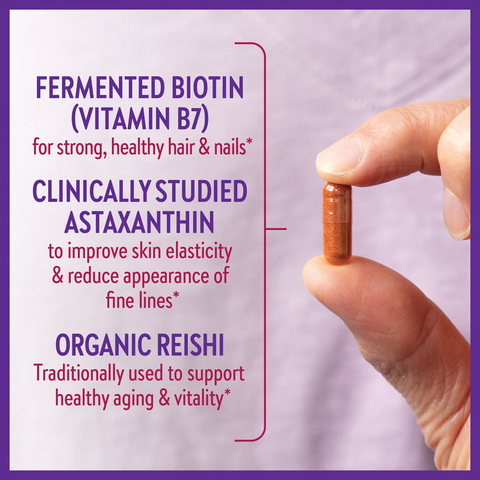 A hand holding a brown capsule against a pink background. Text highlights benefits: "Fermented Biotin (Vitamin B7)" for hair and nails, "Clinically Studied Astaxanthin" for skin elasticity, "Organic Reishi" for healthy aging and vitality.