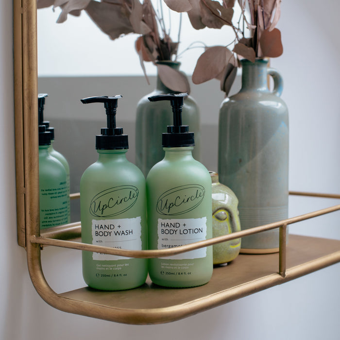 Two green bottles labeled UpCircle Hand + Body Wash and Hand + Body Lotion with Bergamot Water, featuring black pump dispensers, rest on a gold shelf. The citrus-scented lotion adds zest, while muted green vases and dried leaves in the background embrace vegan skincare elegance.